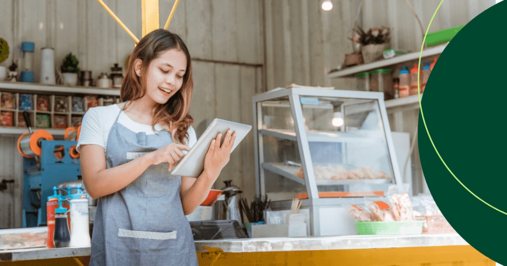 A young female bakery shop owner smiles while using a tablet at her counter, with a bakery display case and shelves of ingredients in the background.