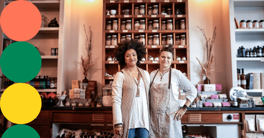 Two women business owners standing together in their specialty shop, surrounded by shelves stocked with jars and packaged goods.