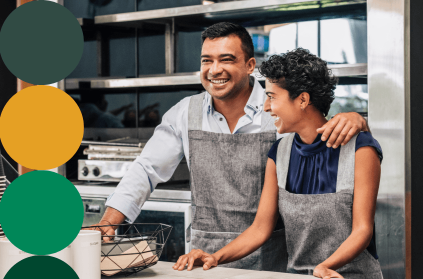Two smiling small business owners wearing gray aprons standing together in a commercial kitchen