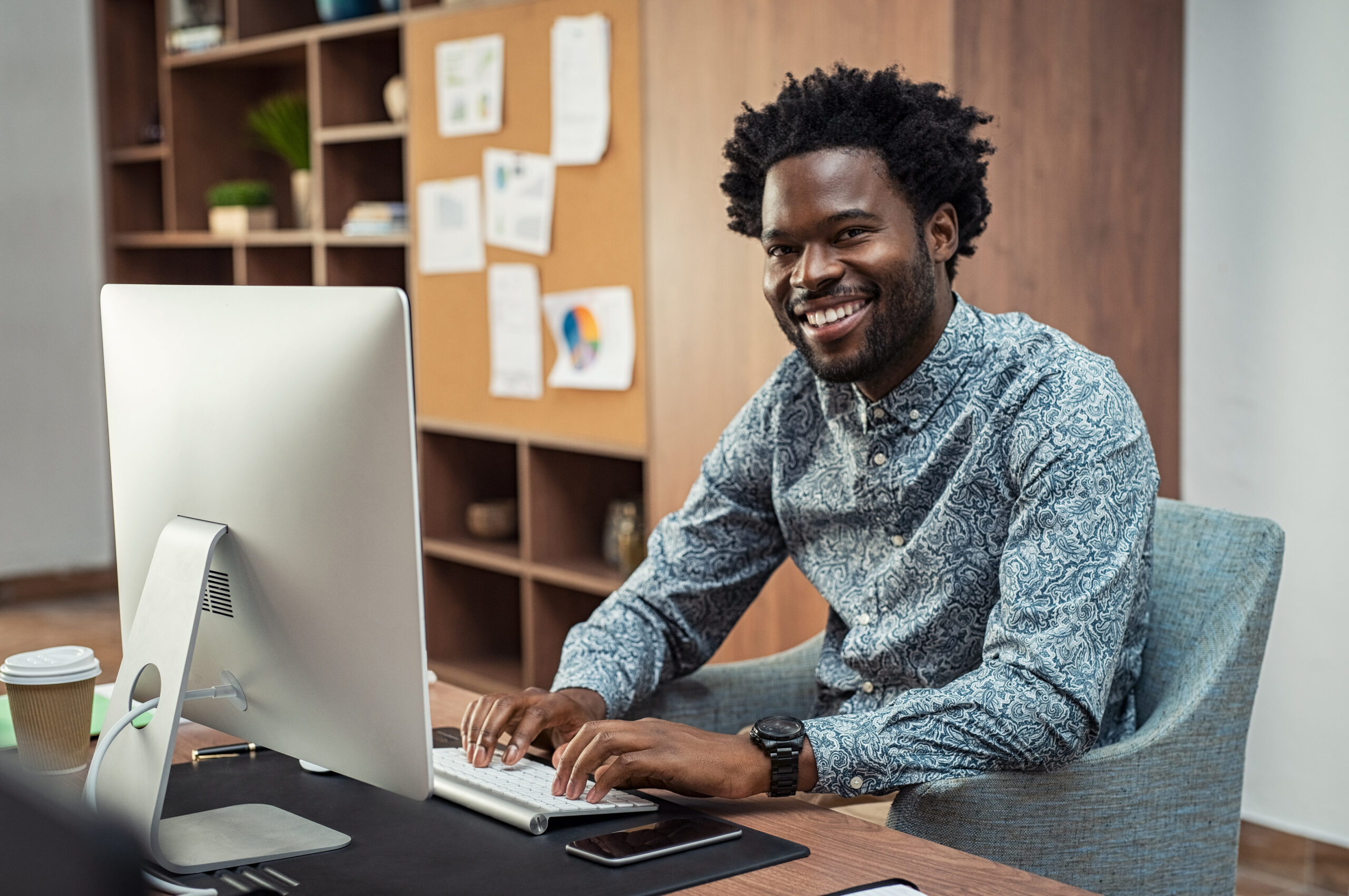 Successful african american businessman typing on desktop computer at modern office. Happy man using computer for work and looking at camera. Portrait of creative business man feeling successful after receiving approval mail from client.