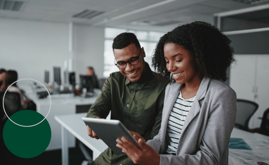 Two colleagues smile while reviewing content on a tablet together in a modern open-plan office. Decorative green circles appear in the lower left corner.