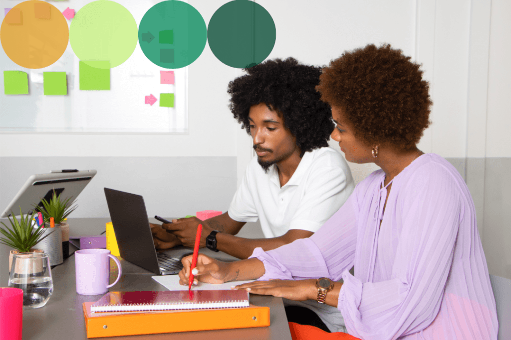 Two colleagues collaborate at a laptop in a bright workspace, with one taking notes while reviewing work together.