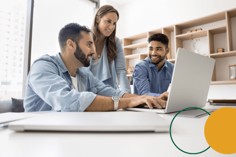 Three colleagues collaborating around a laptop in a bright office workspace, smiling and engaged in discussion.