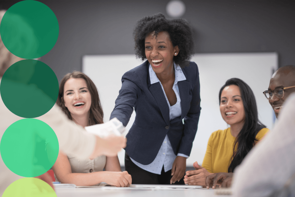 A smiling professional leads an energetic team meeting, passing documents across a conference table to engaged colleagues.