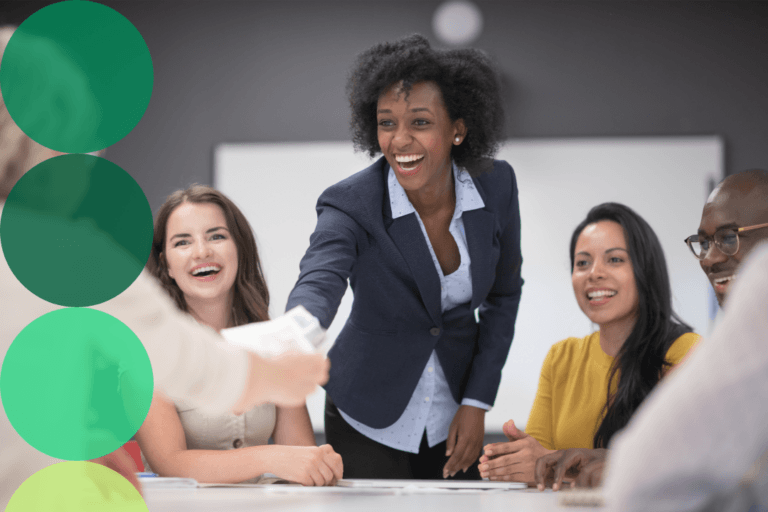 A smiling professional leads an energetic team meeting, passing documents across a conference table to engaged colleagues.
