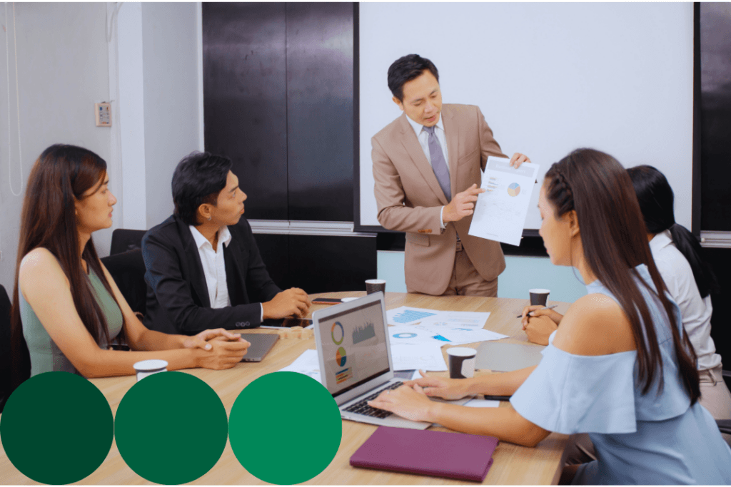 A presenter shares a data report with a team gathered around a conference table, with charts visible on a laptop and printed documents spread across the table.