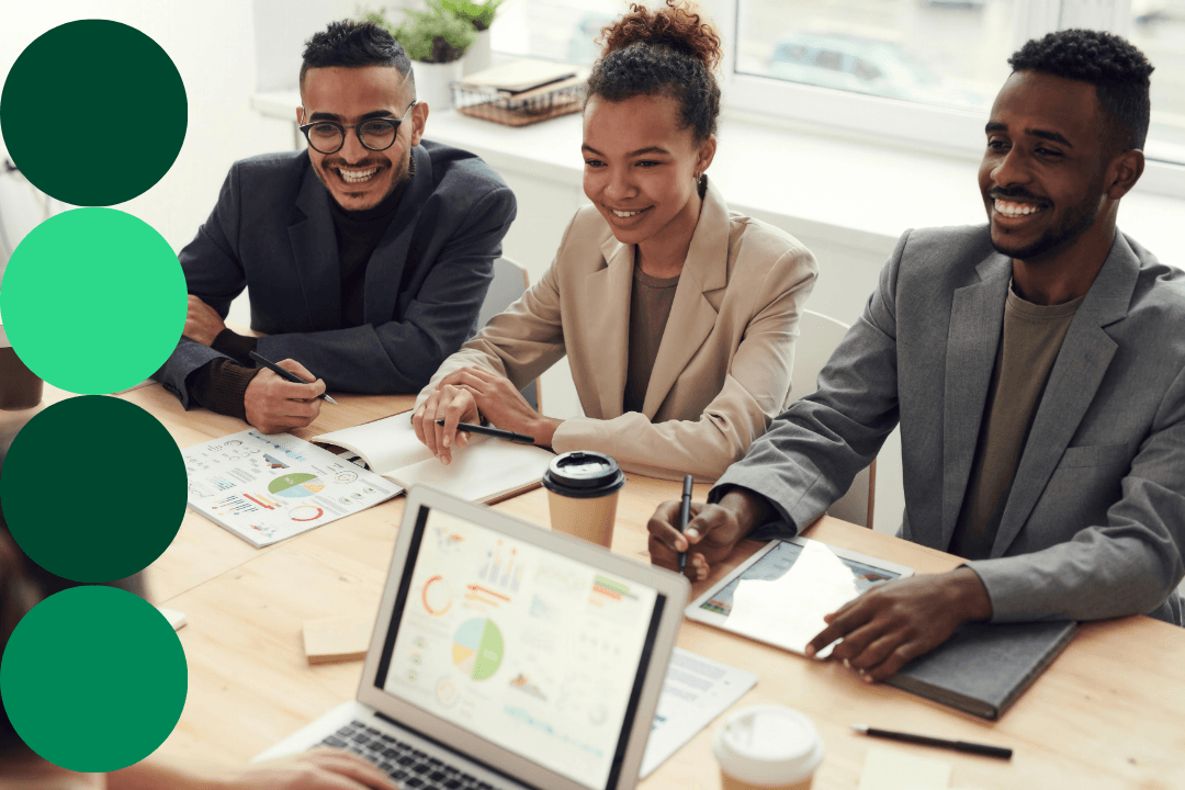 Three business professionals collaborating around a table with data charts, a laptop, and a tablet in a bright office setting.