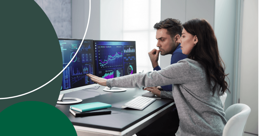 Two procurement professionals analyzing data dashboards on dual monitors, with charts and analytics visualizations displayed on screen.