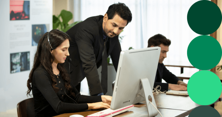 A procurement professional reviews data on a desktop monitor with a colleague looking on in a modern office setting