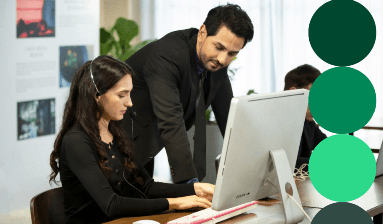 A procurement professional reviews data on a desktop monitor with a colleague looking on in a modern office setting