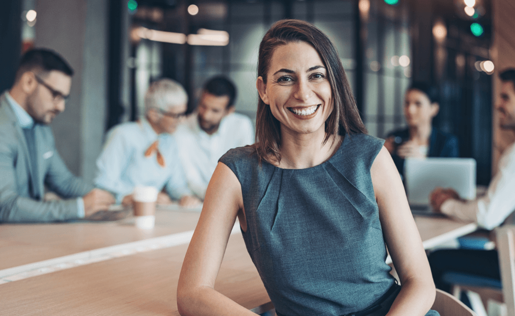 Business professional smiling in a meeting room with colleagues collaborating in the background