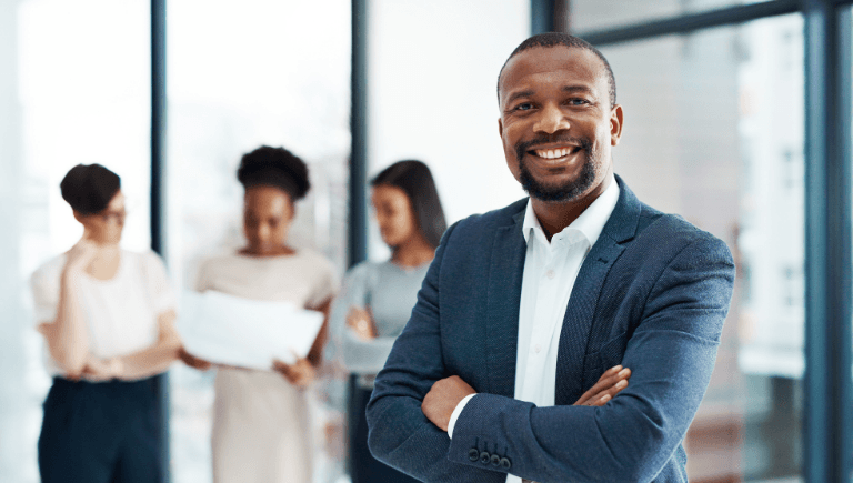 Finance professional smiling with arms crossed, colleagues collaborating in the background