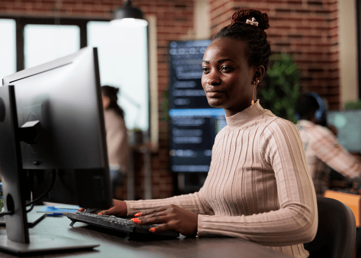 Woman with braided updo working at a desktop computer in a modern office with exposed brick walls.