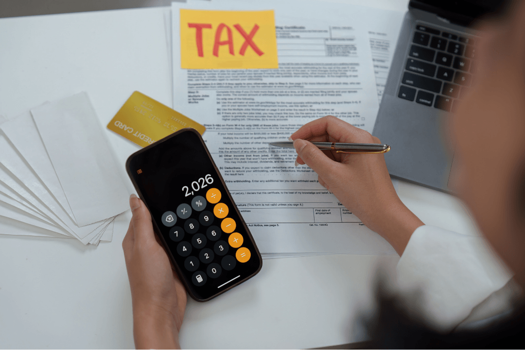 Person filling out a tax withholding form by hand next to a calculator, credit card, and laptop, with a yellow TAX sticky note on the desk.