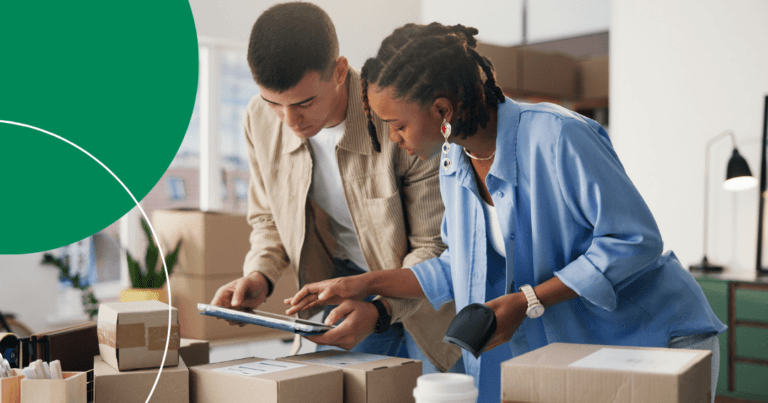 Two coworkers scanning and reviewing shipping boxes together using a tablet and barcode scanner in a warehouse office.