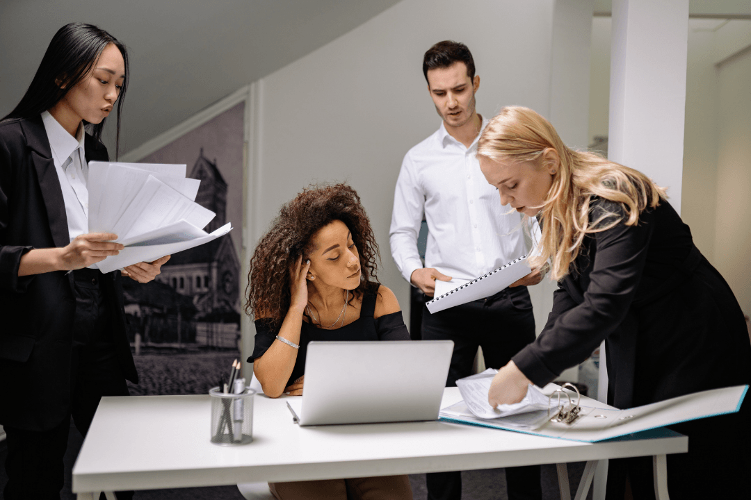 A stressed woman at a laptop surrounded by three colleagues holding papers and documents during a busy office moment.