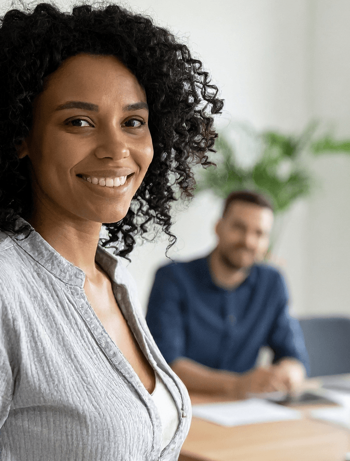 Woman in an office setting smiling with a colleague in the background.