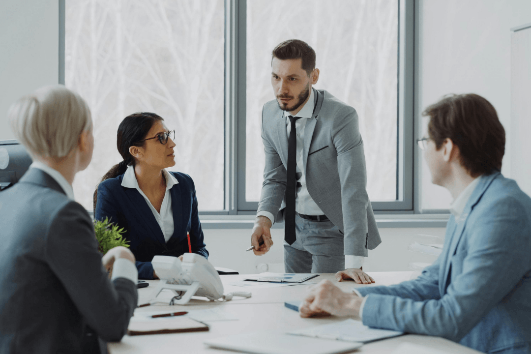 A man standing and speaking to three colleagues seated around a conference table during a business meeting.